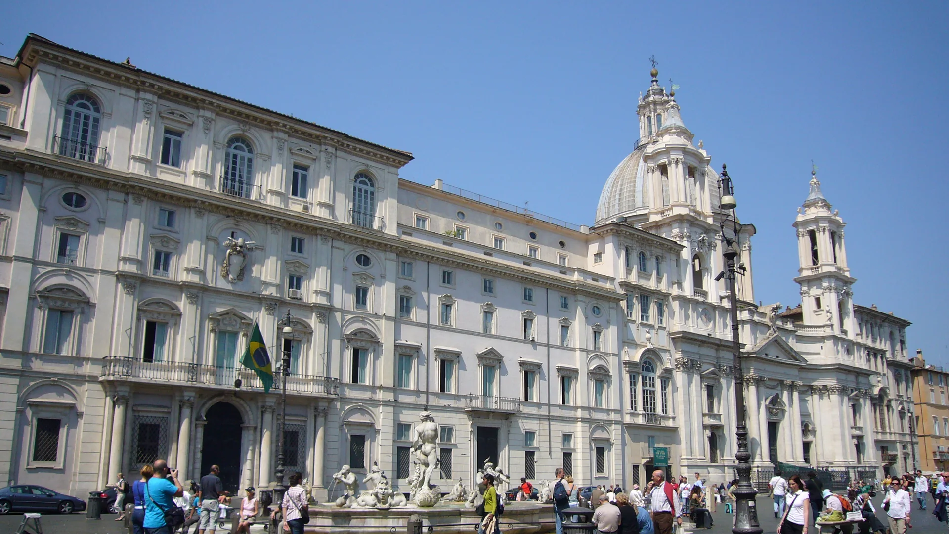 The cream front of Palazzo Pamphilj on Piazza Navona, with the dome and bell tower of Sant'Agnese in Agone rising beside it under a blue sky.