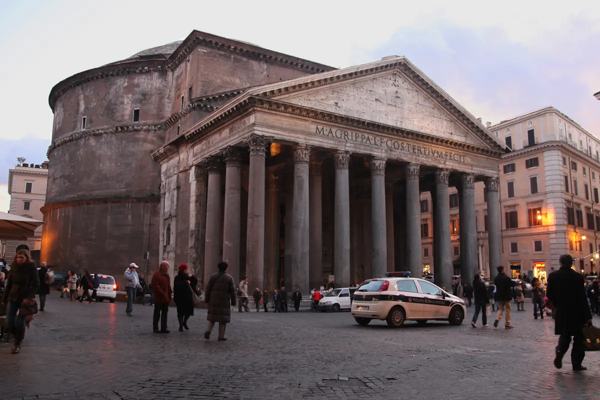 The portico and drum of the Pantheon in the blue light of dusk, with lit windows in the buildings beside it.
