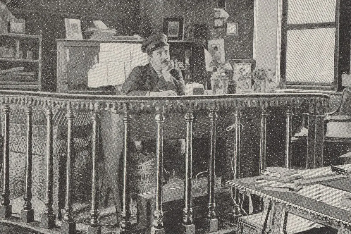 An early printed photograph of Giacomo Puccini in a cap, seated at a desk behind a turned wooden balustrade